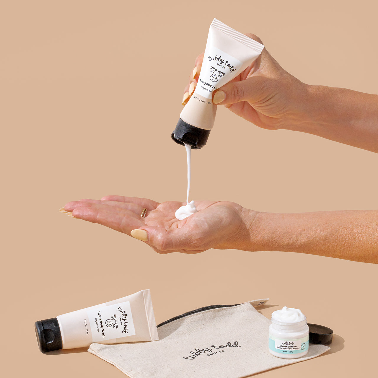 A hand squeezing the fragrance-free 2oz Everyday Lotion tubes on the other hand, and the fragrance-free 2oz Hair + Body Wash tube, 1oz All Over Ointment jar, and the canvas travel bag lying on the beige color background.